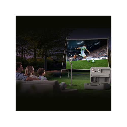 A portable projector set shown in a dark outdoor living room scene, with a family sitting on a couch watching a soccer game on a large screen. The projector and its carrying case are visible in the foreground, along with a soccer ball on the floor.